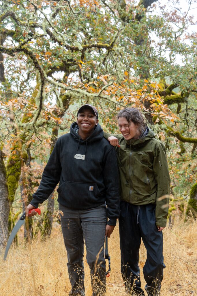 Two people stand in front of a forest of trees at Riddell Preserve. They are both smiling and wear work gloves. 