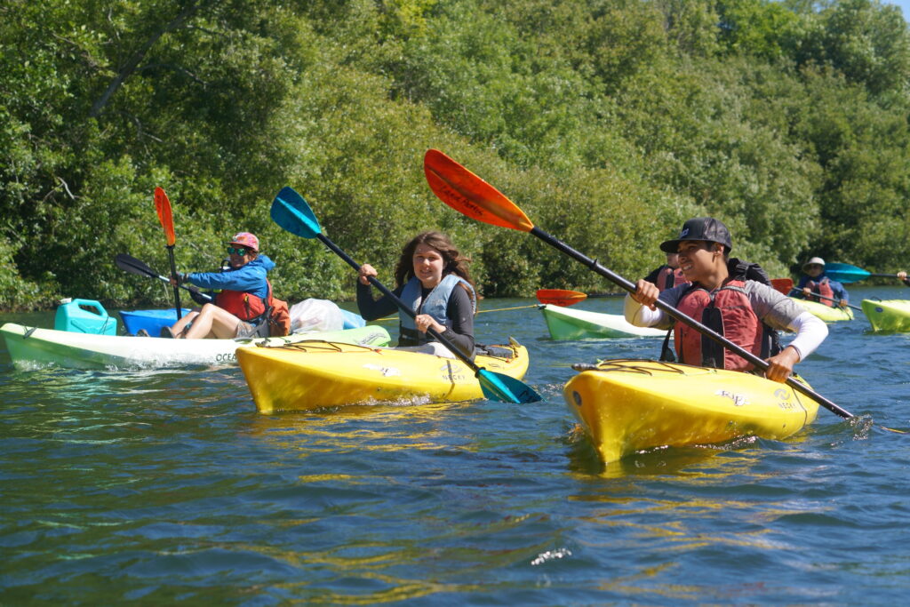 Two teens smile while they are kayaking down the Russian River on a sunny day. They are both wearing flotation vests for safety and are accompanied by an adult chaperon. 