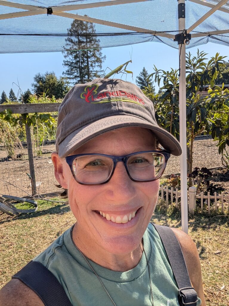 A photo of Mich at Bayer Farm. They are wearing a LandPaths baseball camp, smiling, and a praying mantis is sitting on top of the hat. 